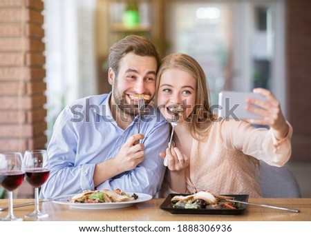 Silly millennial couple having fun during dinner, taking selfie at cozy urban cafe. Pretty lady and her boyfriend making self portrait on smartphone during lunch. Lifestyles and relationships concept