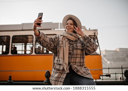Charming woman takes selfie on yellow tram background outside. Happy girl in hat and checkered coat holds phone and shows peace sign outdoors.