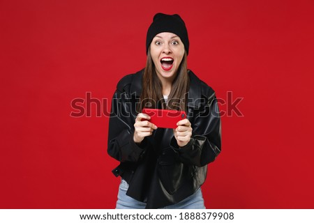Excited surprised amazed young brunette woman 20s in basic casual black leather jacket white t-shirt hat playing game on mobile cell phone isolated on bright red colour background studio portrait