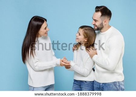 Side view of cheerful young parents mom dad with child kid daughter teen girl in basic white sweaters hugging holding hands isolated on blue background studio portrait. Family day parenthood concept