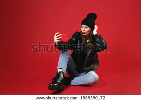 Full length of pretty young brunette woman 20s wearing black leather jacket white t-shirt hat sitting doing selfie shot on mobile phone making duck face isolated on red background studio portrait