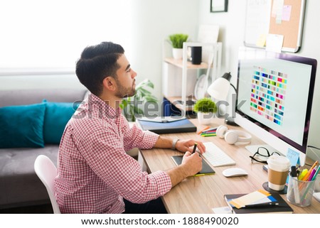 Side view of a latin man in his 20s working as an illustrator that is at his desk designing and working on a new logo or illustration for a client 