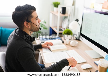 Handsome young man with a headset is employed as an interpreter. Guy in his 20s sitting at his desk is working on translating a writing piece 