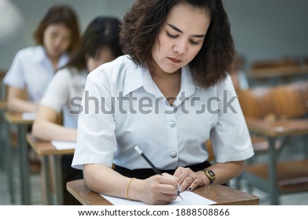 Selective focus of the teenage college students sit on lecture chairs do final examination and write on examination paper answer sheets in the classroom. University students in uniform in classroom.