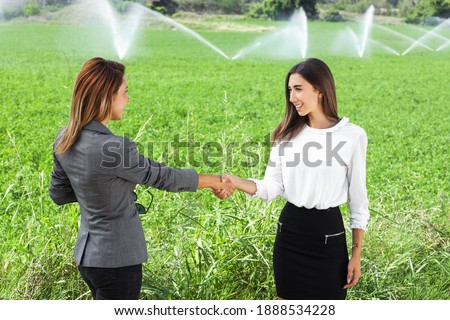 Business women shaking hands in a green field with agriculture irrigation system. Water sprinklers in the background.