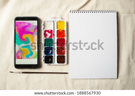 Set of watercolor paints, scetchbook, paintbrush and a smartphone with picture for reference.