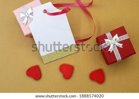 A gift tag next to a red gift box and three red hearts on a yellow background. The concept of greeting and holiday discounts . Price tag sale tag address label.
