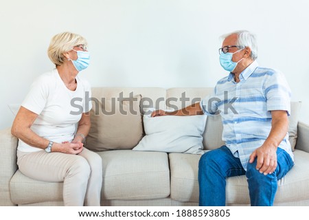 Two Elderly friends sitting in social distance wearing medical face mask and talking on the sofa, preventing covid 19 coronavirus pandemic infection spread. Social distancing on sofa at home