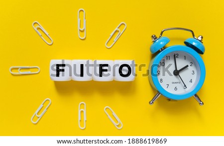 Finance and economics concept. On a yellow background, a blue alarm clock, paper clips and white cubes on which the text is written - FIFO