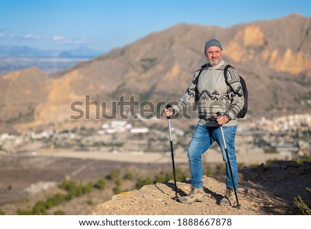 An elderly hiker with a beard in the mountains of southern Spain. It's winter and the man has a backpack and a sweater. In the background are mountains in the sunshine.