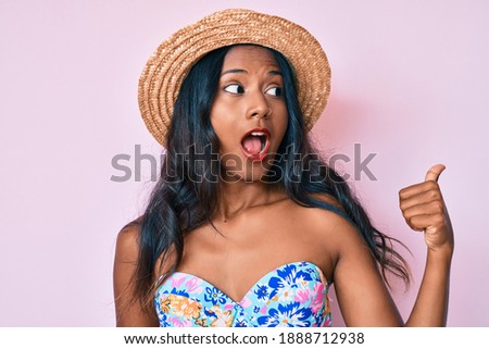 Young indian girl wearing bikini and summer hat smiling with happy face looking and pointing to the side with thumb up. 