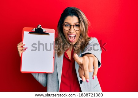 Beautiful brunette woman holding clipboard with blank space smiling and laughing hard out loud because funny crazy joke. 