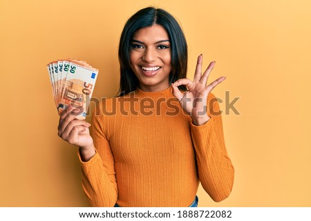 Young latin transsexual transgender woman holding bunch of 50 euro banknotes doing ok sign with fingers, smiling friendly gesturing excellent symbol 