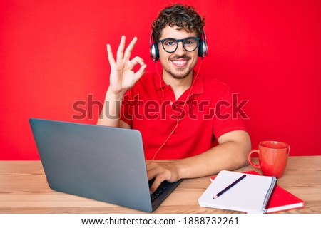 Young caucasian man with curly hair working at the office drinking a cup of coffee doing ok sign with fingers, smiling friendly gesturing excellent symbol 