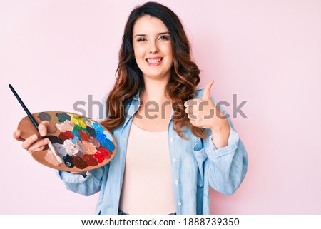 Young beautiful brunette woman holding paintbrush and palette smiling happy and positive, thumb up doing excellent and approval sign 