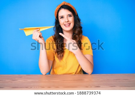 Young beautiful brunette woman holding paper airplane smiling happy pointing with hand and finger 
