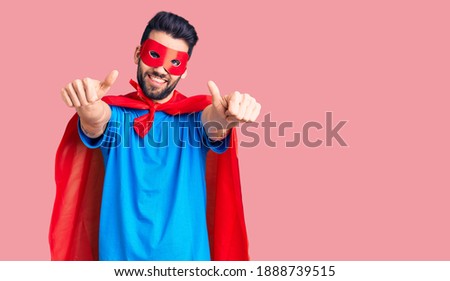 Young handsome man with beard wearing super hero costume approving doing positive gesture with hand, thumbs up smiling and happy for success. winner gesture. 