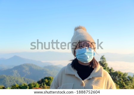 Woman in medical mask traveler standing at mountain with morning fog background and looking at camera.