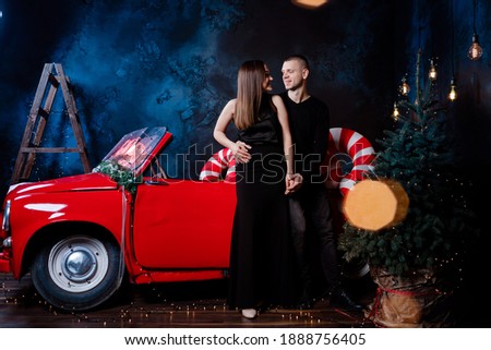 Young happy couple man and woman in love in Christmas are hugging near a red retro car with New Year's gifts. Kiss, girl, happiness, quarantine Christmas celebration, holiday