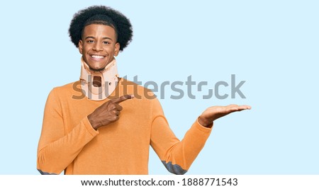African american man with afro hair wearing cervical neck collar amazed and smiling to the camera while presenting with hand and pointing with finger. 