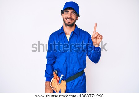 Handsome young man with curly hair and bear weaing handyman uniform showing and pointing up with finger number one while smiling confident and happy. 