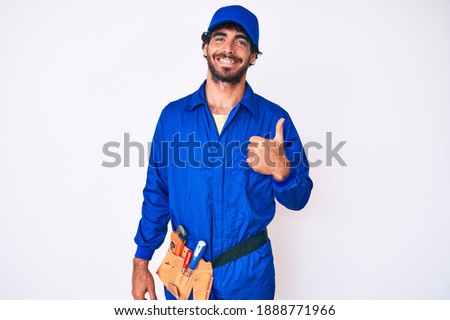 Handsome young man with curly hair and bear weaing handyman uniform doing happy thumbs up gesture with hand. approving expression looking at the camera showing success. 