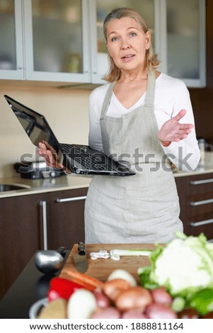 mature woman in the kitchen preparing food and holding laptop