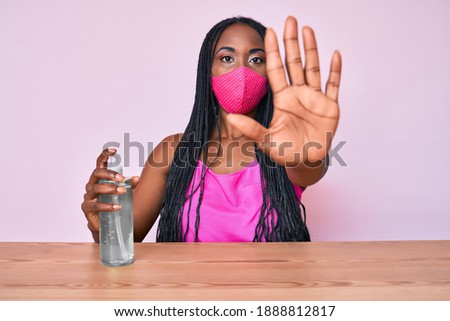 African american woman with braids wearing medical mask holding hand sanitizer gel with open hand doing stop sign with serious and confident expression, defense gesture 