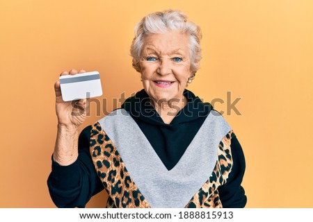 Senior grey-haired woman holding credit card looking positive and happy standing and smiling with a confident smile showing teeth 