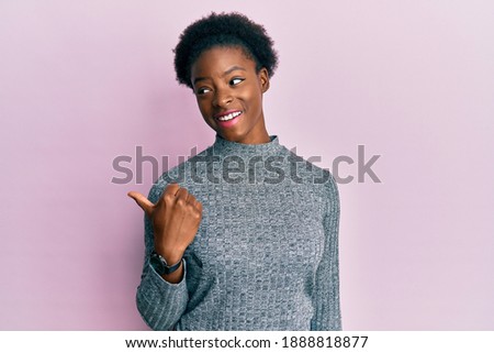 Young african american girl wearing casual clothes smiling with happy face looking and pointing to the side with thumb up. 