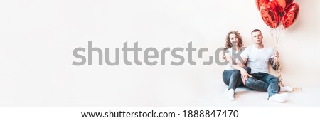 Guy and the girl are sitting near the wall and holding red balloons. Enamored man and woman nearby on Valentine's Day. Banner with copy space for valentine's day. Heterosexual traditional couple