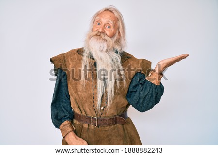 Old senior man with grey hair and long beard wearing viking traditional costume smiling cheerful presenting and pointing with palm of hand looking at the camera. 
