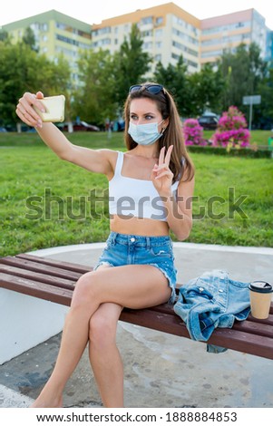 female woman in city summer in medical mask, sits on bench pictures smartphone, online recording on Internet, application phone, video call white topic casual clothes denim jacket, cup of coffee tea