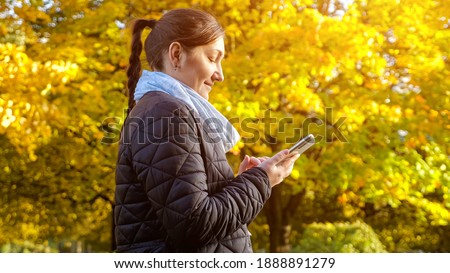 young woman in a black jacket and a gray scarf is typing a message on the phone against the background of yellowed trees copyspace