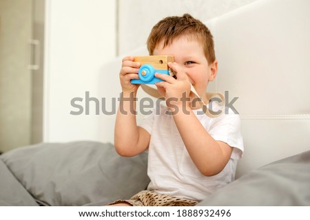 A small cheerful child plays with a wooden environmental toy at home on a white bed.The boy takes pictures with a blue camera.