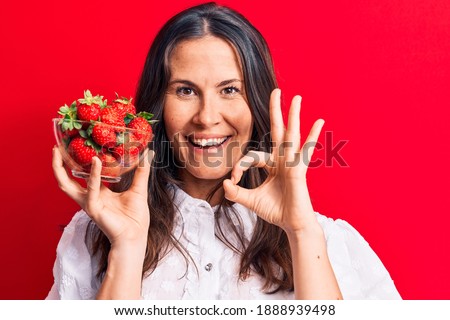 Young beautiful brunette woman holding bowl of heallthy strawberries over red background doing ok sign with fingers, smiling friendly gesturing excellent symbol
