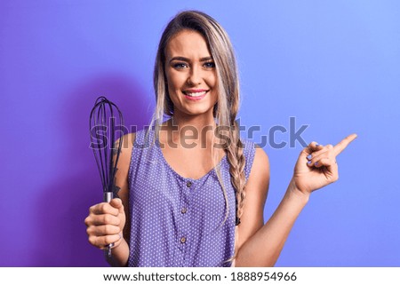 Young beautiful blonde woman cooking holding whisk over isolated purple background smiling happy pointing with hand and finger to the side