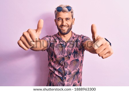 Handsome blond man on vacation wearing casual shirt and sunglasses over pink background approving doing positive gesture with hand, thumbs up smiling and happy for success. Winner gesture.