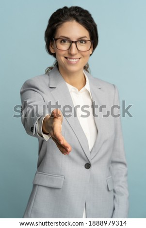 Studio portrait of friendly hospitable cheerful business woman or office worker wear blazer giving hand to handshake, hostess greeting guests, looking at camera, blue background. Focus on hand.  Royalty-Free Stock Photo #1888979314