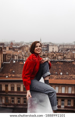 Pretty lady in red sweater and jeans sincerely smiles on terrace with European city view. Beautiful woman sits on fence on balcony.