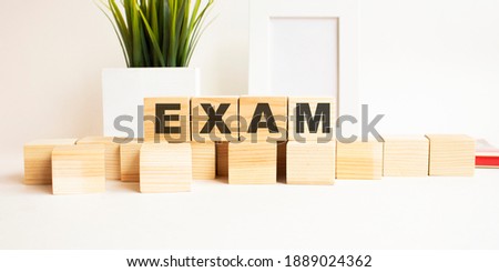 Wooden cubes with letters on a white table. The word is EXAM. White background with photo frame, house plant.