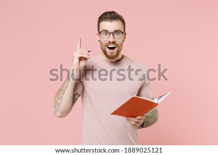Amazed young bearded tattooed man in pastel casual t-shirt, eyeglasses hold notebook pen, point index finger up with great new idea isolated on pink background studio. Tattoo translate life is fight