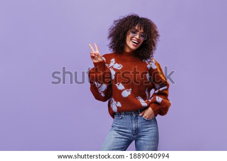 Curly brunette dark-skinned woman smiles sincerely and shows peace sign. Joyful girl poses on isolated purple background.