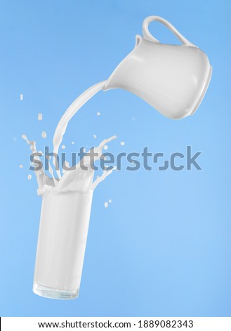 Pouring milk from jug into glass cup with splashing on blue background Royalty-Free Stock Photo #1889082343