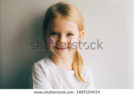 Pretty smilling blue-eyed blonde little  girl with pink hearts on her cheeks on a white, grey background. Child and valentines day. Love. Pink hearts on a face.Happy girl.