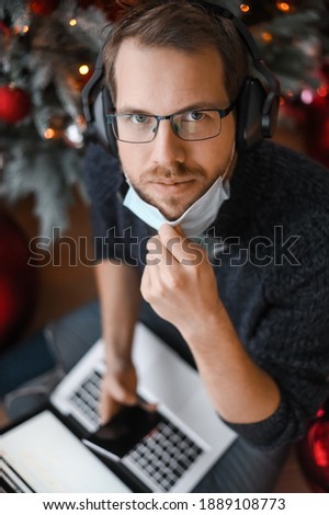Portrait of nerd man with Christmas tree on the background holding phone smartphone above the laptop during the covid-19 pandemic with face mask at home office