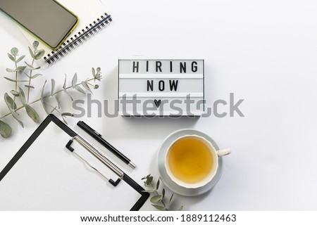 Creative flat lay overhead view composition with text Hiring Now on the lightbox, clipboard with white blank paper, cup of herbal tea. Concept of hiring and recruitment. 
