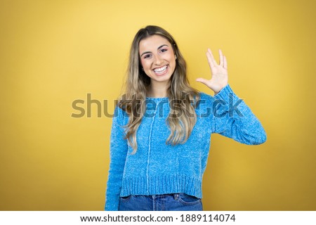Pretty blonde woman with long hair standing over yellow background doing hand symbol