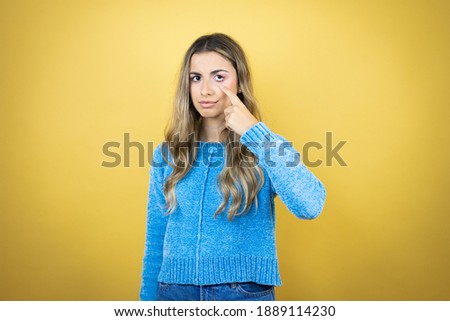 Pretty blonde woman with long hair standing over yellow background Pointing to the eye watching you gesture, suspicious expression