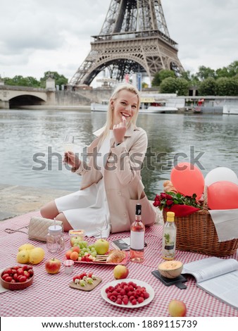 A beautiful blonde in a white dress sits on a checkered tablecloth on the banks of the Seine and holds a glass of wine. Summer French picnic on the background of the Eiffel Tower in Paris.
